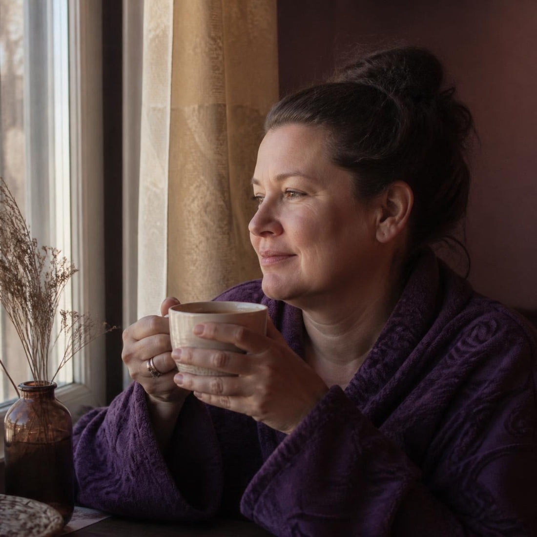A woman in a purple wrap holds a warm cup of tea with both hands, gazing peacefully out a sunlit window beside dried botanicals.