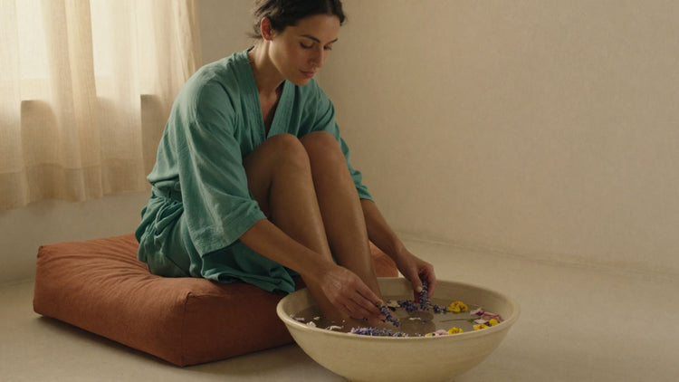 A woman in a soft teal robe sits on the floor, feet submerged in a wide cream bowl of milky water, hands gently dipping in — surrounded by rose petals, lavender sprigs, and yellow blooms in warm evening light.