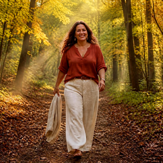 A middle-aged woman with long wavy dark brown hair walks confidently along a sunlit dirt forest path, mid-stride and smiling genuinely. She
wears a terracotta linen blouse, wide-leg cream pants, flat sandals, a delicate necklace, and small earrings, with a cream linen jacket draped loosely
in one hand. Warm golden light streams through trees transitioning from green to deep gold and amber, fallen autumn leaves covering the path beneath her
feet. Energizing and free — a real woman coming back to herself.