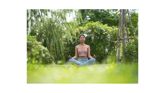 Young woman seated cross-legged on grass in a lush garden, eyes closed in meditation, hands in mudra, surrounded by soft green foliage and warm natural light.