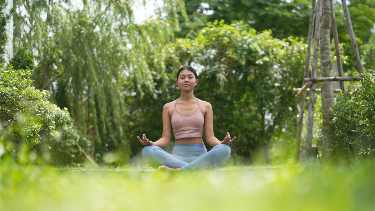 Young woman seated cross-legged on grass in a lush garden, eyes closed in meditation, hands in mudra, surrounded by soft green foliage and warm natural light.